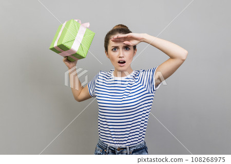Portrait of shocked surprised woman wearing striped T-shirt holding far away with open mouth and hand over forehead, holding present box. Indoor studio shot isolated on gray background. Portrait of shocked surprised woman wearing striped T-shirt holding far away with open mouth and hand over forehead, holding present box. Indoor studio shot isolated on gray background. 108268975