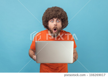 Portrait of astonished shocked man with Afro hairstyle wearing orange T-shirt standing holding laptop and looking at camera with open mouth and big eyes. Indoor studio shot isolated on blue background 108269017