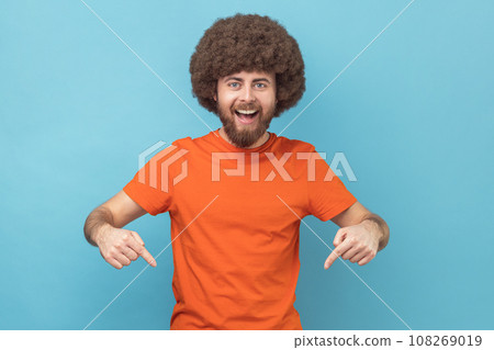 Portrait of excited satisfied man with Afro hairstyle wearing orange T-shirt pointing down with fore fingers, looking at camera with toothy smile. Indoor studio shot isolated on blue background. Portrait of excited satisfied man with Afro hairstyle wearing orange T-shirt pointing down with fore fingers, looking at camera with toothy smile. Indoor studio shot isolated on blue background. 108269019