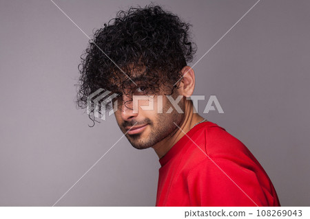 Portrait of attractive handsome young adult bearded man with curly hair, wearing red jumper, looking at camera, showing his hairdo. Indoor studio shot isolated on gray background. 108269043