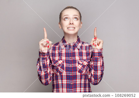 Portrait of hopeful woman with bun hairstyle standing looking pointing up with clenched teeth, copy space for advertisement, wearing checkered shirt. Indoor studio shot isolated on gray background. 108269052