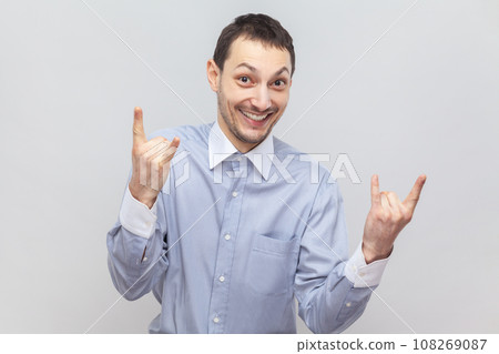Portrait of funny positive smiling man showing rock and roll gesture, expressing positive emotions, being happy at concert, wearing light blue shirt. Indoor studio shot isolated on gray background. 108269087