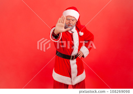 Portrait of serious elderly man with gray beard wearing santa claus costume showing stop ban gesture with palm, sign of prohibition, with hand. Indoor studio shot isolated on red background. 108269098