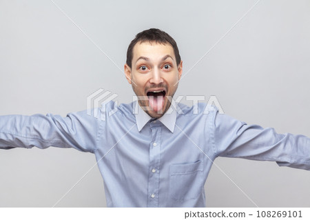 Portrait of funny crazy childish man standing looking at camera, keeps mouth widely open, showing tongue out, wearing light blue shirt. Indoor studio shot isolated on gray background. 108269101