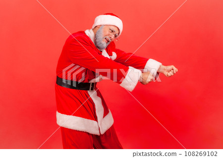 Elderly man with gray beard in santa claus costume pretending to pull invisible rope, concept of hard working, striving and efforts to achievements. Indoor studio shot isolated on red background. 108269102