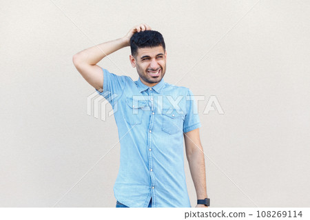 Portrait of puzzled confused pensive young adult man wearing denim shirt standing with hand on head, thinking, trying to remember something. Indoor studio shot isolated on gray background. 108269114