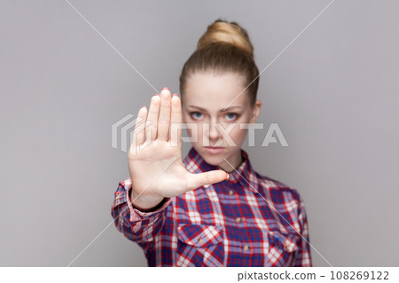 Portrait of serious bossy woman with bun hairstyle showing prohibition gesture, shows stop palm sign, looking at camera, wearing checkered shirt. Indoor studio shot isolated on gray background. Portrait of serious bossy woman with bun hairstyle showing prohibition gesture, shows stop palm sign, looking at camera, wearing checkered shirt. Indoor studio shot isolated on gray background. 108269122