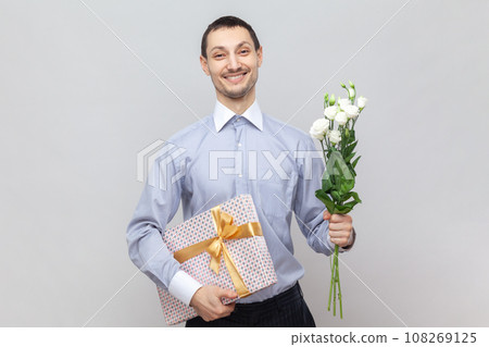 Smiling happy positive man standing with present box and bouquet of flowers, congratulate her girlfriend with birthday, wearing light blue shirt. Indoor studio shot isolated on gray background. 108269125