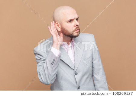 Portrait of bald man overhearing, listening intently to secret information, private talk, holding hand near ear to hear better, wearing gray jacket. Indoor studio shot isolated on brown background. Portrait of bald man overhearing, listening intently to secret information, private talk, holding hand near ear to hear better, wearing gray jacket. Indoor studio shot isolated on brown background. 108269127