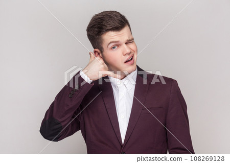 Portrait of handsome young man makes call me gesture, winking his eyes, flirting with beautiful girl, wearing violet suit and white shirt. Indoor studio shot isolated on grey background. Portrait of handsome young man makes call me gesture, winking his eyes, flirting with beautiful girl, wearing violet suit and white shirt. Indoor studio shot isolated on grey background. 108269128