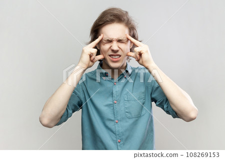 Portrait of sick ill unhealthy young man standing massaging his temples, suffering headache, frowning face, keeps eyes closed, wearing blue shirt. Indoor studio shot isolated on gray background. Portrait of sick ill unhealthy young man standing massaging his temples, suffering headache, frowning face, keeps eyes closed, wearing blue shirt. Indoor studio shot isolated on gray background. 108269153