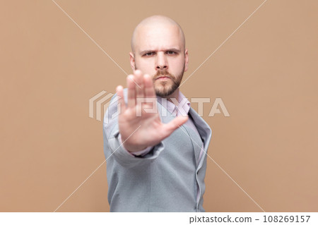 Portrait of bald bearded man does stop sign with palm, demonstrates prohibition and restriction, refuses something, says no, wearing gray jacket. Indoor studio shot isolated on brown background. 108269157