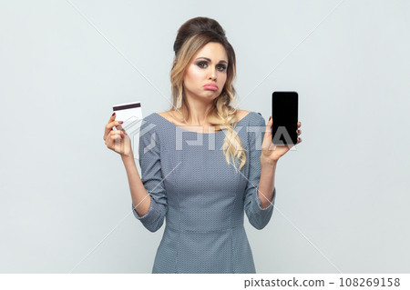 Portrait of sad upset disappointed blonde woman holding smart phone with empty display with copy space and credit card, wearing grey elegant dress. Indoor studio shot isolated on gray background. 108269158