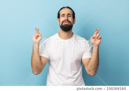 Portrait of man with beard wearing white T-shirt crossed prays for miracle anticipates for important results stands with eyes closed wants to win. Indoor studio shot isolated on blue background. 108269160