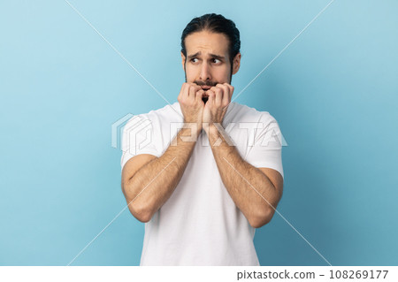 Troubles and worries. Portrait of man with beard wearing white T-shirt biting nails, terrified about problems, suffering phobia, anxiety disorder. Indoor studio shot isolated on blue background. 108269177