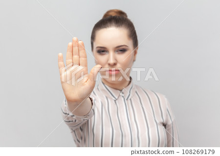 Portrait of young adult strict serious woman wearing striped shirt standing showing prohibition gesture with her palm, asking to stop. Indoor studio shot isolated on gray background. Portrait of young adult strict serious woman wearing striped shirt standing showing prohibition gesture with her palm, asking to stop. Indoor studio shot isolated on gray background. 108269179