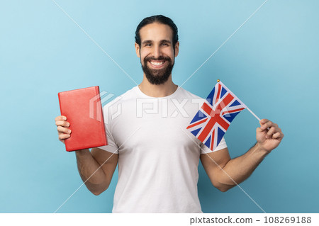 Portrait of smiling positive man with beard wearing white T-shirt holding book and Great Britain flag, education, learning English language. Indoor studio shot isolated on blue background. Portrait of smiling positive man with beard wearing white T-shirt holding book and Great Britain flag, education, learning English language. Indoor studio shot isolated on blue background. 108269188
