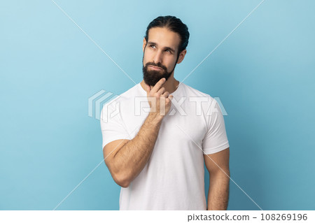 Portrait of thoughtful handsome man with beard wearing white T-shirt holding his chin and pondering idea, confused not sure about solution. Indoor studio shot isolated on blue background. 108269196