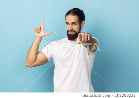 Portrait of frustrated man with beard wearing white T-shirt showing looser gesture and pointing at camera, looking with grumpy face. Indoor studio shot isolated on blue background. 108269232