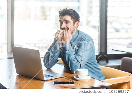 Nervous worried young man freelancer in blue jeans shirt working on laptop, looking at display, biting his finger nails, waiting for interview results. Indoor shot near big window, cafe background. Nervous worried young man freelancer in blue jeans shirt working on laptop, looking at display, biting his finger nails, waiting for interview results. Indoor shot near big window, cafe background. 108269237