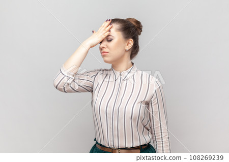 Portrait of stressed depressed attractive woman wearing striped shirt standing showing facepalm gesture, forgot something important. Indoor studio shot isolated on gray background. Portrait of stressed depressed attractive woman wearing striped shirt standing showing facepalm gesture, forgot something important. Indoor studio shot isolated on gray background. 108269239