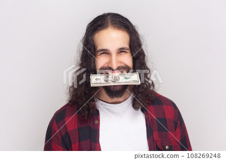 Portrait of rich funny positive happy bearded man with long curly hair in checkered red shirt holding dollar banknotes in his mouth, looking at camera. Indoor studio shot isolated on gray background. Portrait of rich funny positive happy bearded man with long curly hair in checkered red shirt holding dollar banknotes in his mouth, looking at camera. Indoor studio shot isolated on gray background. 108269248