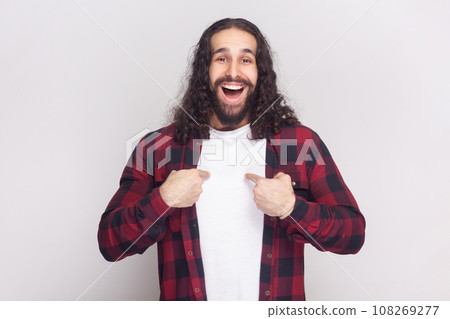 Pleased smiling bearded man with long curly hair in checkered red shirt points at himself, looks with proud happy expression boasts of his success. Indoor studio shot isolated on gray background. Pleased smiling bearded man with long curly hair in checkered red shirt points at himself, looks with proud happy expression boasts of his success. Indoor studio shot isolated on gray background. 108269277