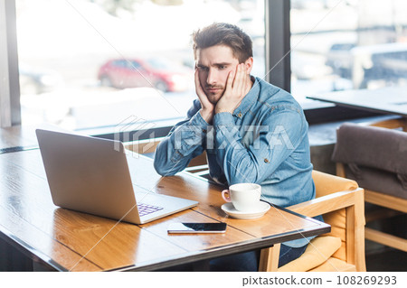 Portrait of upset stressed attractive young man freelancer in blue jeans shirt working on laptop, looking at display, expressing sadness and sorrow. Indoor shot near big window, cafe background. 108269293
