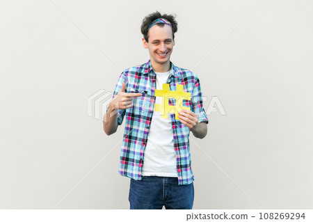 Smiling man holding and pointing at yellow hashtag, setting trending topics on a social media app, wearing blue checkered shirt and headband. Indoor studio shot isolated on gray background. 108269294