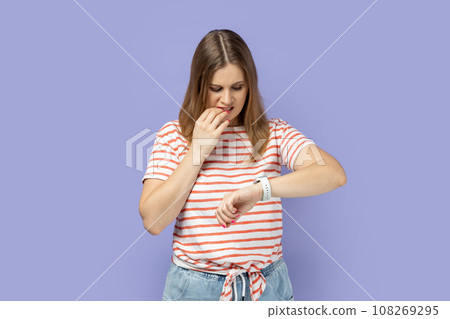 Portrait of nervous blond woman wearing striped T-shirt standing looking at smartwatch and biting her nails, being stressed and worried. Indoor studio shot isolated on purple background. 108269295