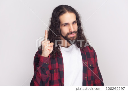 Attention please. Portrait of serious bearded man with long curly hair in checkered red shirt raises finger up, warning, has confident look. Indoor studio shot isolated on gray background. 108269297