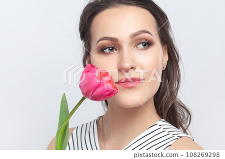 Closeup portrait of romantic dreaming brunette woman standing looking away, holding pink tulip, enjoying aromat of flower, wearing striped dress. Indoor studio shot isolated on gray background. 108269298