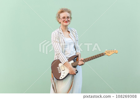 Portrait of beautiful positive woman in white shirt, skirt, and striped blouse with eyeglasses, holding guitar and looking at camera with smile. Indoor studio shot isolated on light green background. Portrait of beautiful positive woman in white shirt, skirt, and striped blouse with eyeglasses, holding guitar and looking at camera with smile. Indoor studio shot isolated on light green background. 108269308
