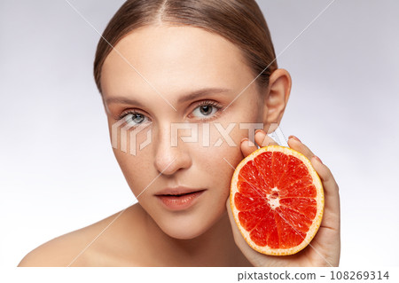 Closeup portrait of beautiful woman with perfect skin model looking at camera holding half of grapefruit. Indoor studio shot isolated over gray background. Closeup portrait of beautiful woman with perfect skin model looking at camera holding half of grapefruit. Indoor studio shot isolated over gray background. 108269314