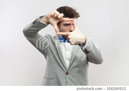 Portrait of focused concentrated bearded man standing looking at camera through frame from fingers, making photo, wearing grey suit and blue bow tie. Indoor studio shot isolated on gray background. 108269322
