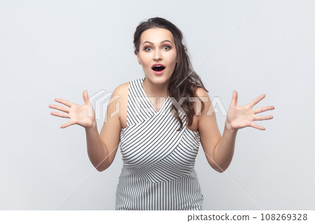 Portrait of woman exclaims happily, keeps palms raised, feels very happy, reacts on awesome news, gets pleasant surprise, wearing striped dress. Indoor studio shot isolated on gray background. Portrait of woman exclaims happily, keeps palms raised, feels very happy, reacts on awesome news, gets pleasant surprise, wearing striped dress. Indoor studio shot isolated on gray background. 108269328