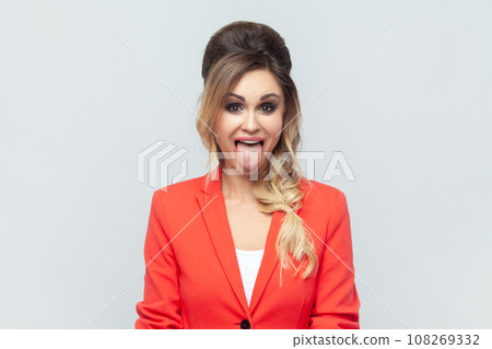 Portrait of funny crazy attractive blonde woman standing sticking tongue out, demonstrates childish behavior, wearing red jacket. Indoor studio shot isolated on gray background. Portrait of funny crazy attractive blonde woman standing sticking tongue out, demonstrates childish behavior, wearing red jacket. Indoor studio shot isolated on gray background. 108269332