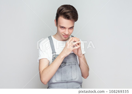 My plan is perfect. Young brunette man schemes something, steepls fingers and looks with cunning expression aside, smiles sly, wearing denim overalls. Indoor studio shot isolated on gray background. 108269335