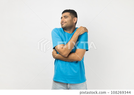 Portrait of happy optimistic smiling unshaven man wearing blue T- shirt standing hugging himself, looks satisfied, expressing happiness. Indoor studio shot isolated on gray background. 108269344