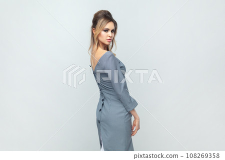Side view portrait of winsome charming blonde woman fashion model, looking at camera, expressing confidence, wearing grey elegant dress. Indoor studio shot isolated on gray background. Side view portrait of winsome charming blonde woman fashion model, looking at camera, expressing confidence, wearing grey elegant dress. Indoor studio shot isolated on gray background. 108269358