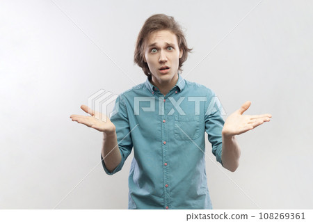 Portrait of unhappy stressed young man standing raised arms and asking, expressing negative emotions, frowning face, wearing blue shirt. Indoor studio shot isolated on gray background. 108269361