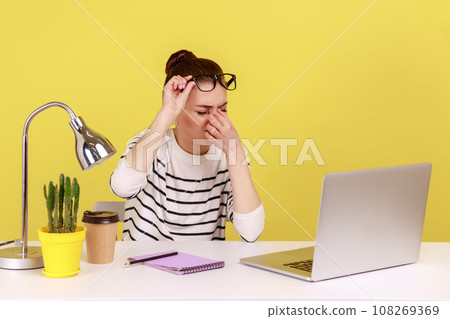 Exhausted stressed woman office worker rubbing tired of laptop work eyes, putting off glasses, sitting workplace, overworking. Indoor studio studio shot isolated on yellow background. Exhausted stressed woman office worker rubbing tired of laptop work eyes, putting off glasses, sitting workplace, overworking. Indoor studio studio shot isolated on yellow background. 108269369