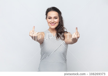 Portrait of brunette woman showing fuck you sign, looking with smile, being vulgar and has quarrel with someone, wearing striped dress. Indoor studio shot isolated on gray background. Portrait of brunette woman showing fuck you sign, looking with smile, being vulgar and has quarrel with someone, wearing striped dress. Indoor studio shot isolated on gray background. 108269373