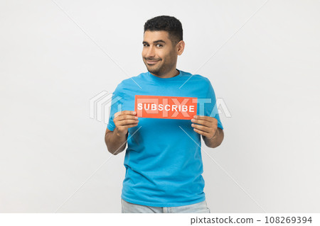 Portrait of smiling optimistic unshaven man in blue T- shirt standing holding card with subscribe inscription, looking at camera with happy expression. Indoor studio shot isolated on gray background. Portrait of smiling optimistic unshaven man in blue T- shirt standing holding card with subscribe inscription, looking at camera with happy expression. Indoor studio shot isolated on gray background. 108269394