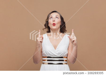 Portrait of surprised astonished middle aged woman with wavy hair standing pointing upwards at advertising area, wearing white dress. Indoor studio shot isolated on light brown background. Portrait of surprised astonished middle aged woman with wavy hair standing pointing upwards at advertising area, wearing white dress. Indoor studio shot isolated on light brown background. 108269395