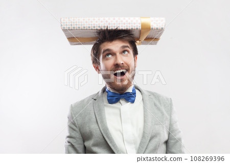 Portrait of overjoyed funny bearded man standing with present box on head, expressing happiness, wearing grey suit and blue bow tie. Indoor studio shot isolated on gray background. Portrait of overjoyed funny bearded man standing with present box on head, expressing happiness, wearing grey suit and blue bow tie. Indoor studio shot isolated on gray background. 108269396