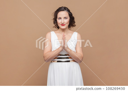 Portrait of smiling happy middle aged woman with wavy hair standing keeps palms together, looking at camera, wearing white dress. Indoor studio shot isolated on light brown background. 108269430