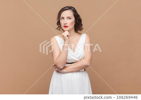 People, facial expressions and thoughts. Portrait of middle aged woman with wavy hair holding her chin, thinking, wearing white dress. Indoor studio shot isolated on light brown background. People, facial expressions and thoughts. Portrait of middle aged woman with wavy hair holding her chin, thinking, wearing white dress. Indoor studio shot isolated on light brown background. 108269446