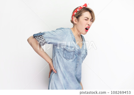 Portrait of sick blonde attractive woman wearing blue denim shirt and red headband standing touching her painful back, frowning face from terrible pain. Indoor studio shot isolated on gray background. 108269453