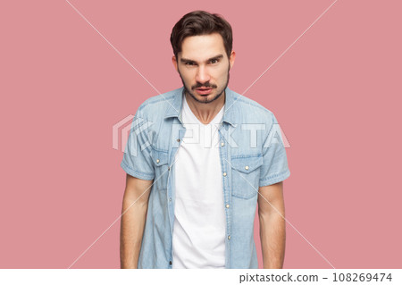 Portrait of bearded man in blue casual style shirt standing frowning, having grumpy angry look, dissatisfied with misbehavior of somebody. Indoor studio shot isolated on pink background. Portrait of bearded man in blue casual style shirt standing frowning, having grumpy angry look, dissatisfied with misbehavior of somebody. Indoor studio shot isolated on pink background. 108269474